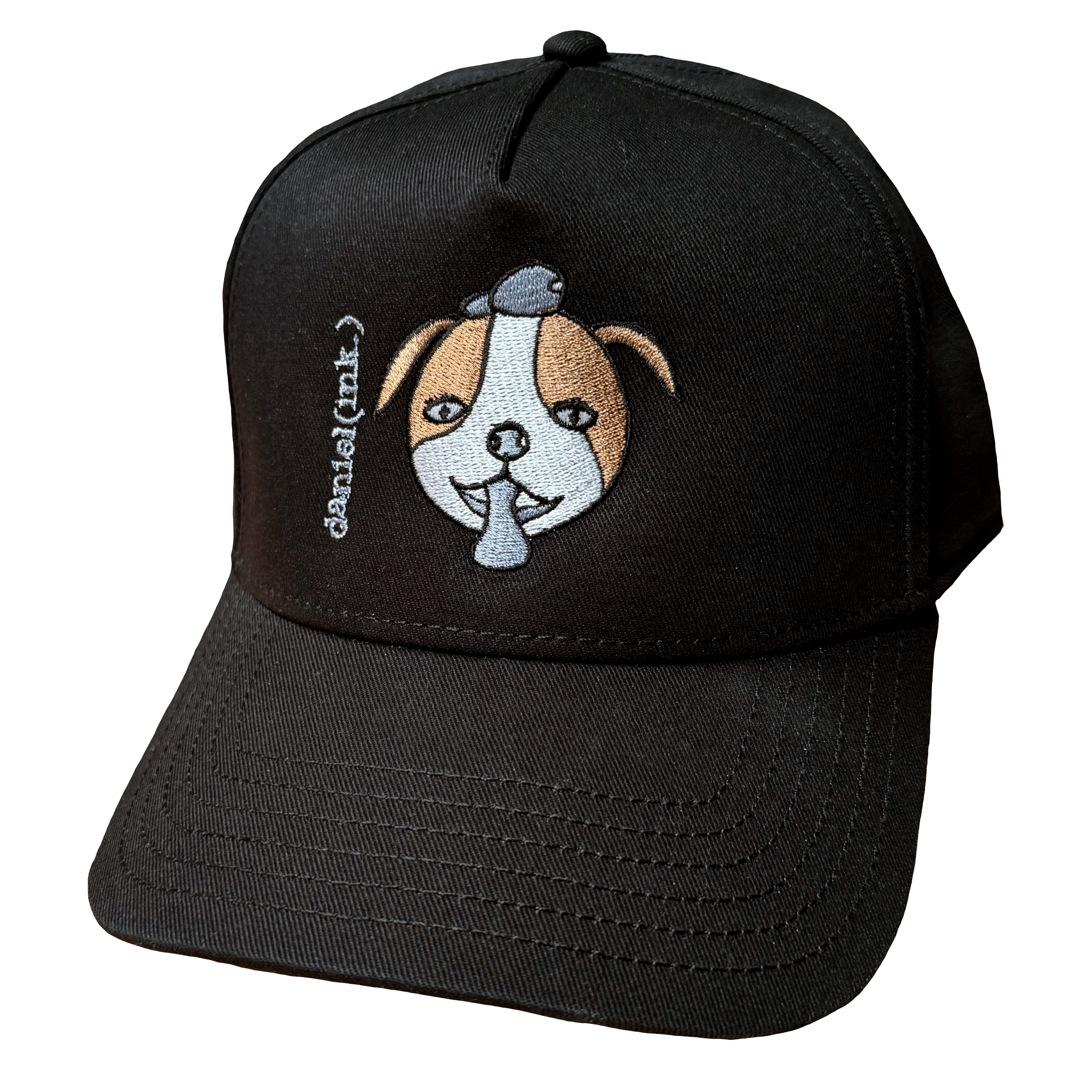 Black snapback cap featuring an embroidered image of a dog's face in brown and white. The dog has a round head, floppy ears, wide eyes, and its tongue is sticking out. The name daniel(ink.) is embroidered in silver thread next to the dog's face.