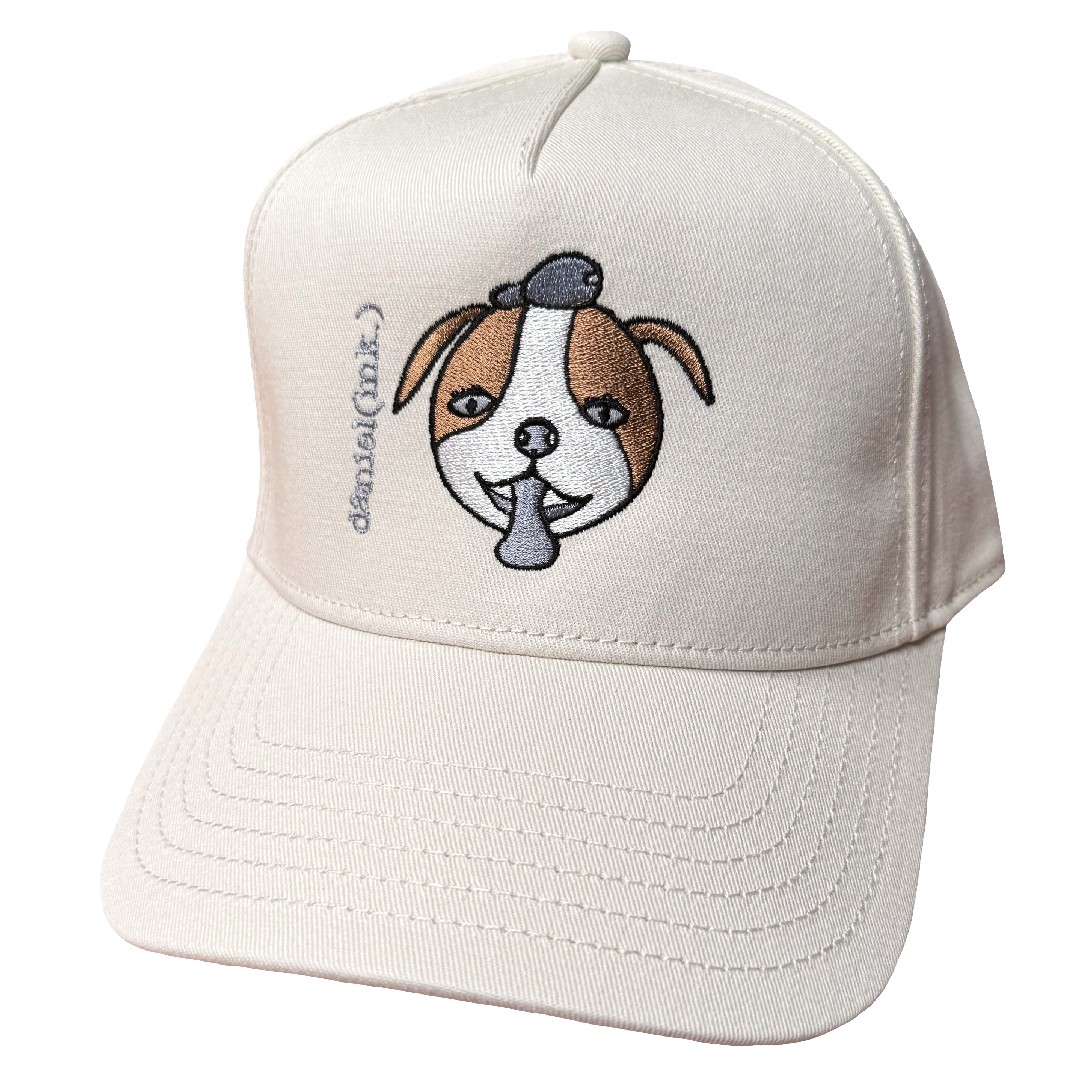 Ecru snapback cap featuring an embroidered image of a dog's face in brown and white. The dog has a round head, floppy ears, wide eyes, and its tongue is sticking out. The name daniel(ink.) is embroidered in silver thread next to the dog's face.